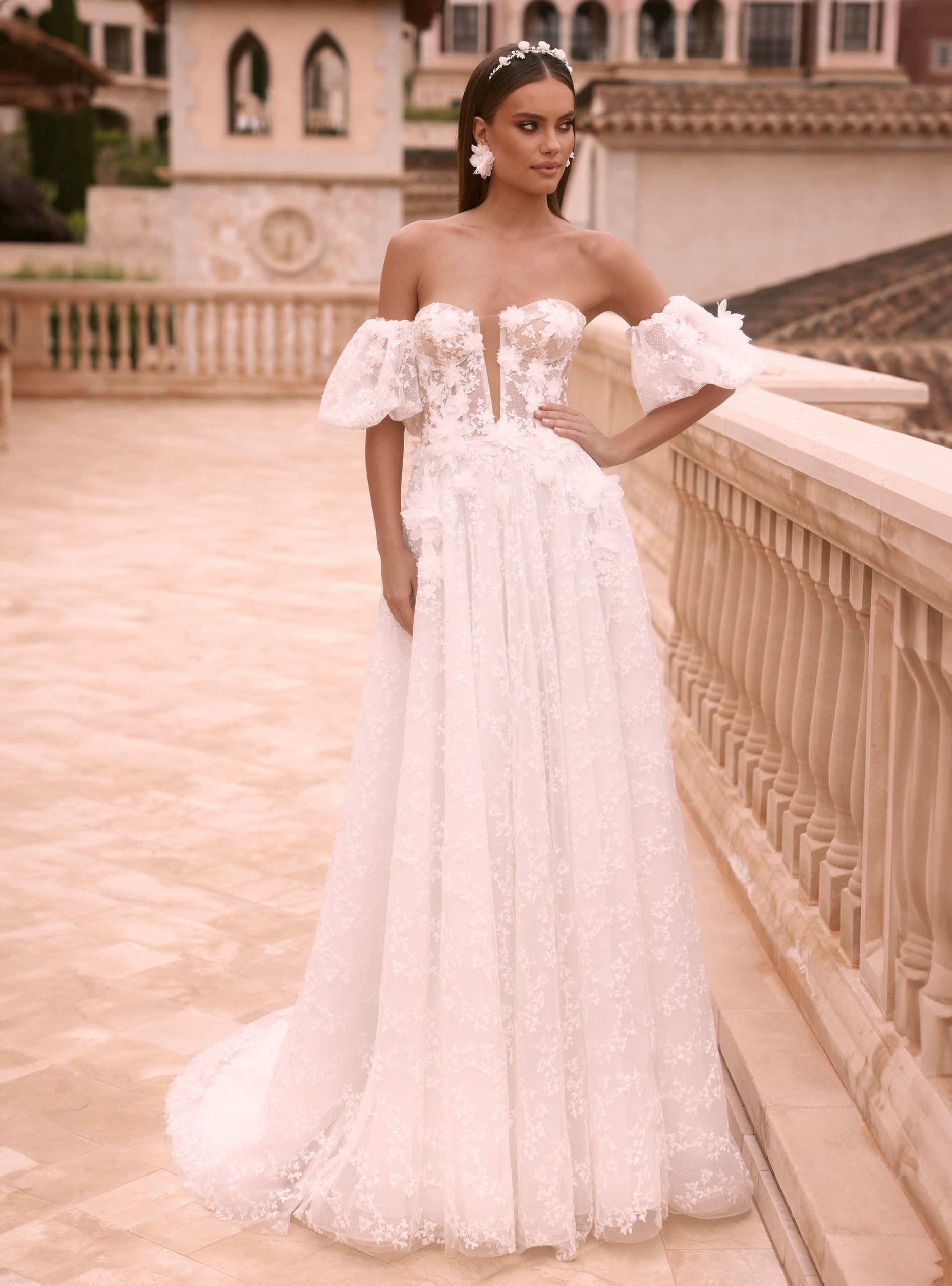 Eine junge Braut trägt ein schulterfreies A-Linien-Corsagenkleid mit Lingerie-Effekt, 3D-Blüten, abnehmbaren Ballonärmeln und langer Schleppe. Das Kleid besteht aus feiner Spitze und gehört zur Kollektion von AMORELY Brautmoden Velbert, in der Nähe von Dortmund