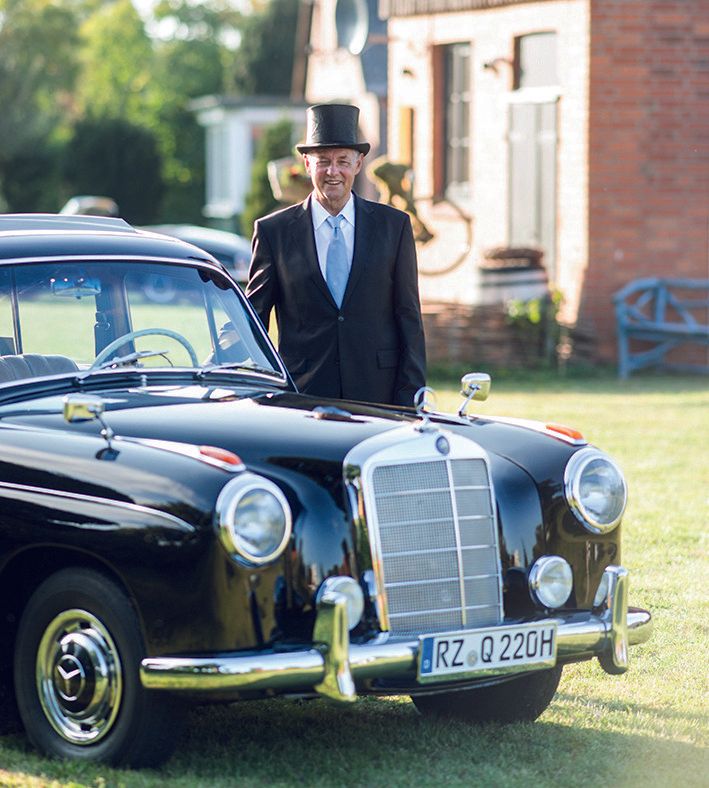 Chauffeur Wolgang Meissner, im schwarzen Anzug mit weißen Hemd und Krawatte sowie einem schwarzen Zylinder, stehend hinter einem schwarzen Oldtimer, Mercedes Ponton. Der Oldtimer steht auf der Wiese vor dem Gebäude der Alten Sägerei, welche man im Hintergrund erkennen kann.