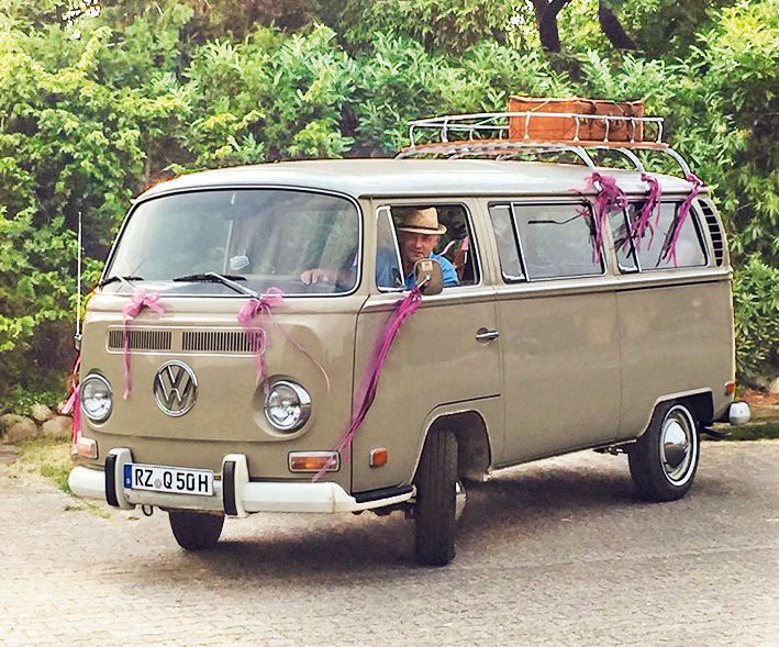 VW Bulli T2 A mit lila Bändern geschmückt. Der Oldtimer steht vor einem Hintergrund aus grünen Blättern. Im Auto sitzt mit einem Strohhut auf dem Kopf der Chaffeur der Alten Sägerei, Wolgang Meissner.