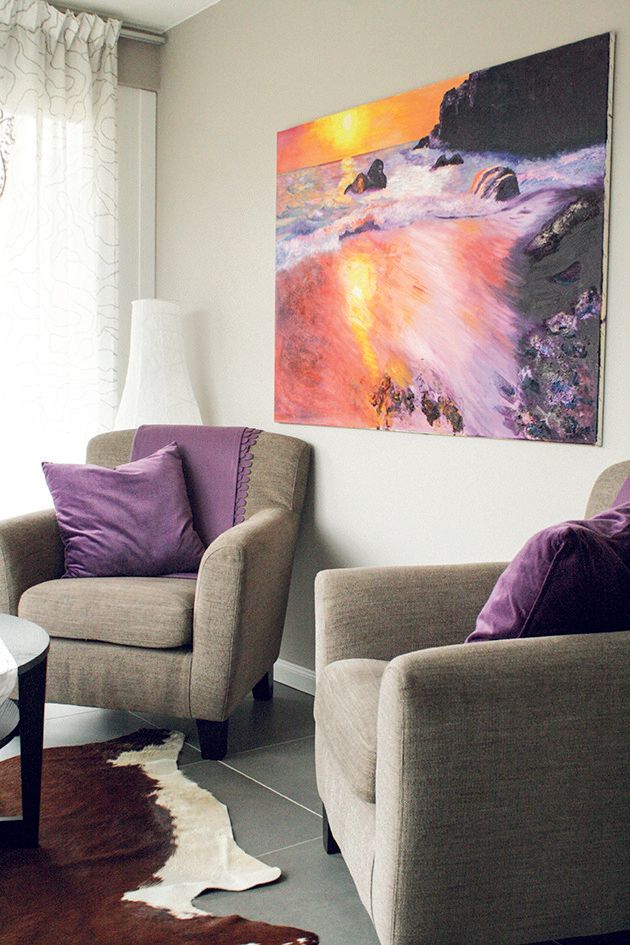 Wohnzimmer in der Ferienwohnung der Alten Sägerei. Auf dem grauen Boden liegt ein Kuhfell, darauf steht ein Tisch. An der rückwärtigen Wand stehen zwei graue Sessel mit jeweils einem lila Kissen drauf. Vor dem großen Fenster hinten links hängen weiße Gardinen. An der rückwärtigen Wand hinten rechts hängt ein großes Bild, welches einen Sonnenuntergang zeigt.