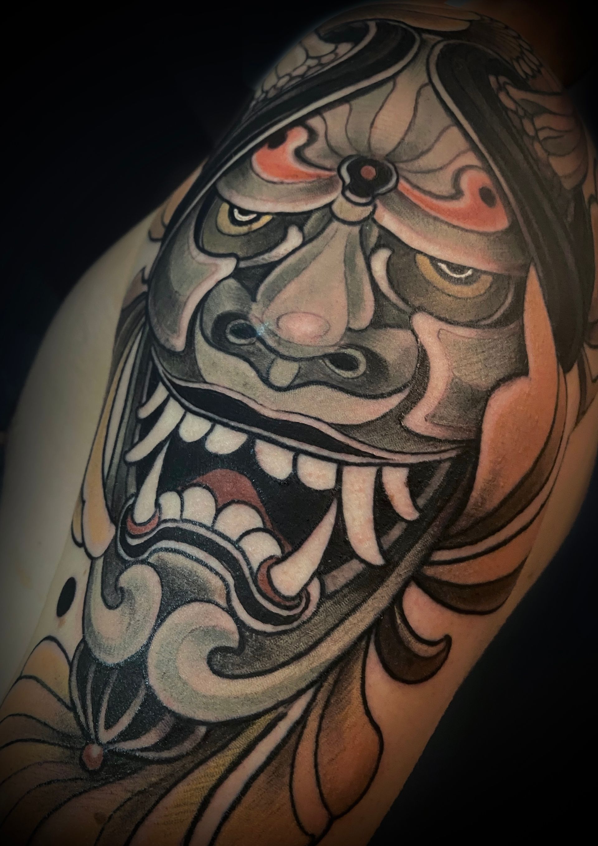 Neo Japanese Hanija Maske Tattoo auf Oberarm umrahmt mit Blätter. Gestochen von Patrick Schneider im Tattoo Studio Berlin.