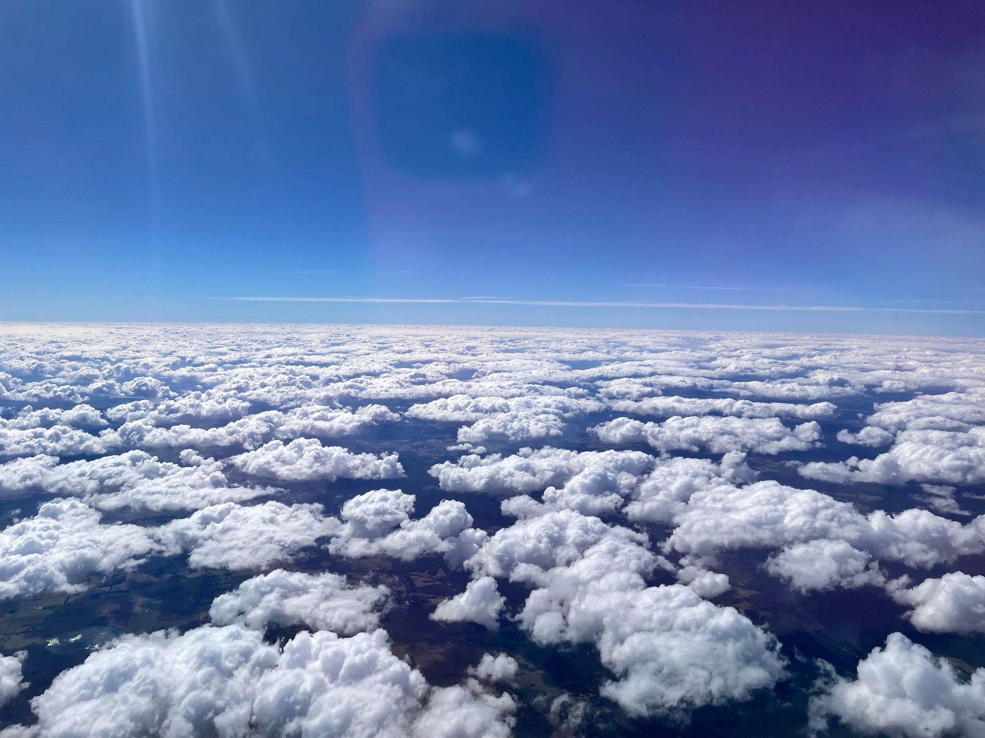 Photo taken from a plane of white fluffy clouds below and a deep blue sky.