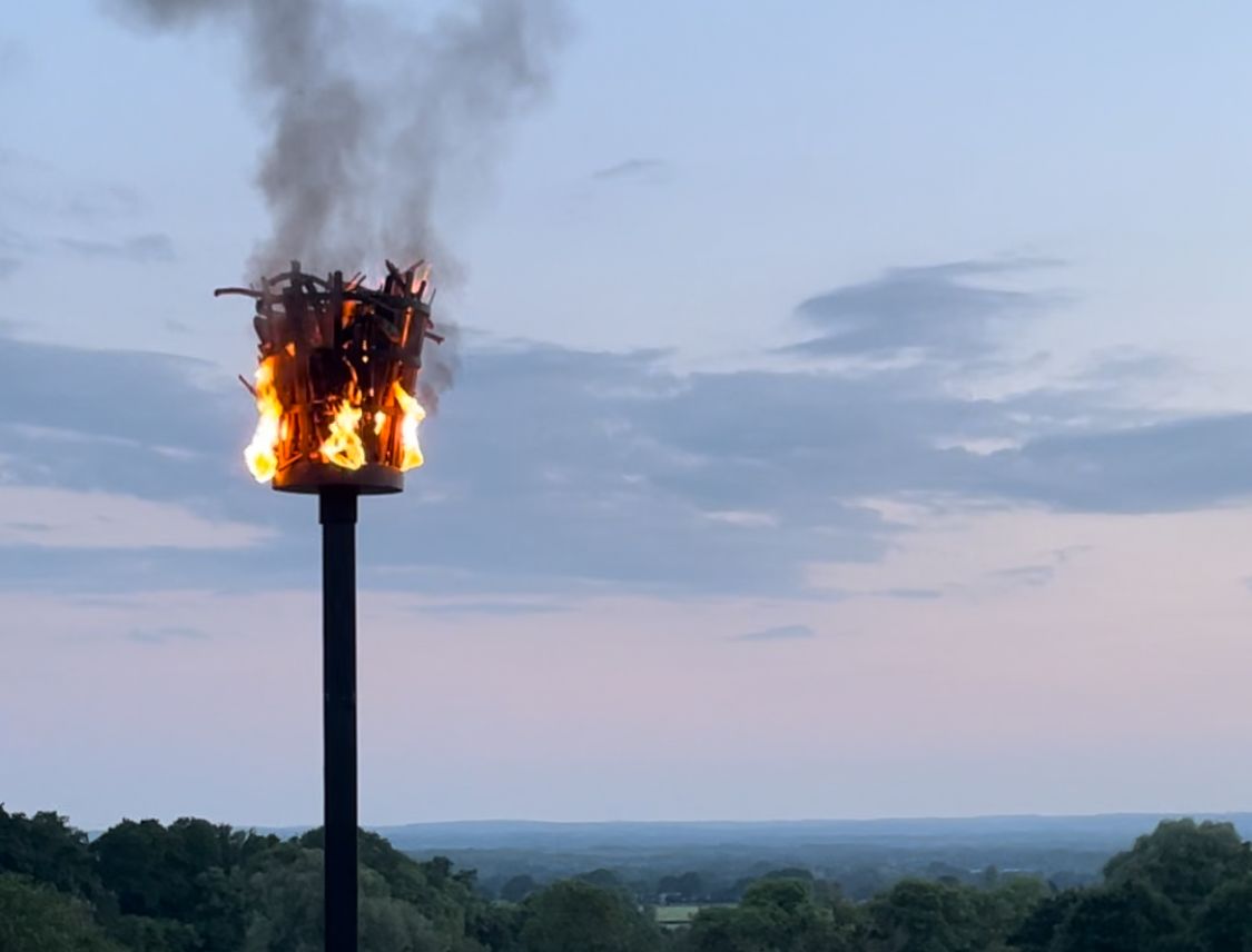 Image of a lit beacon at dusk with a view across green countryside.