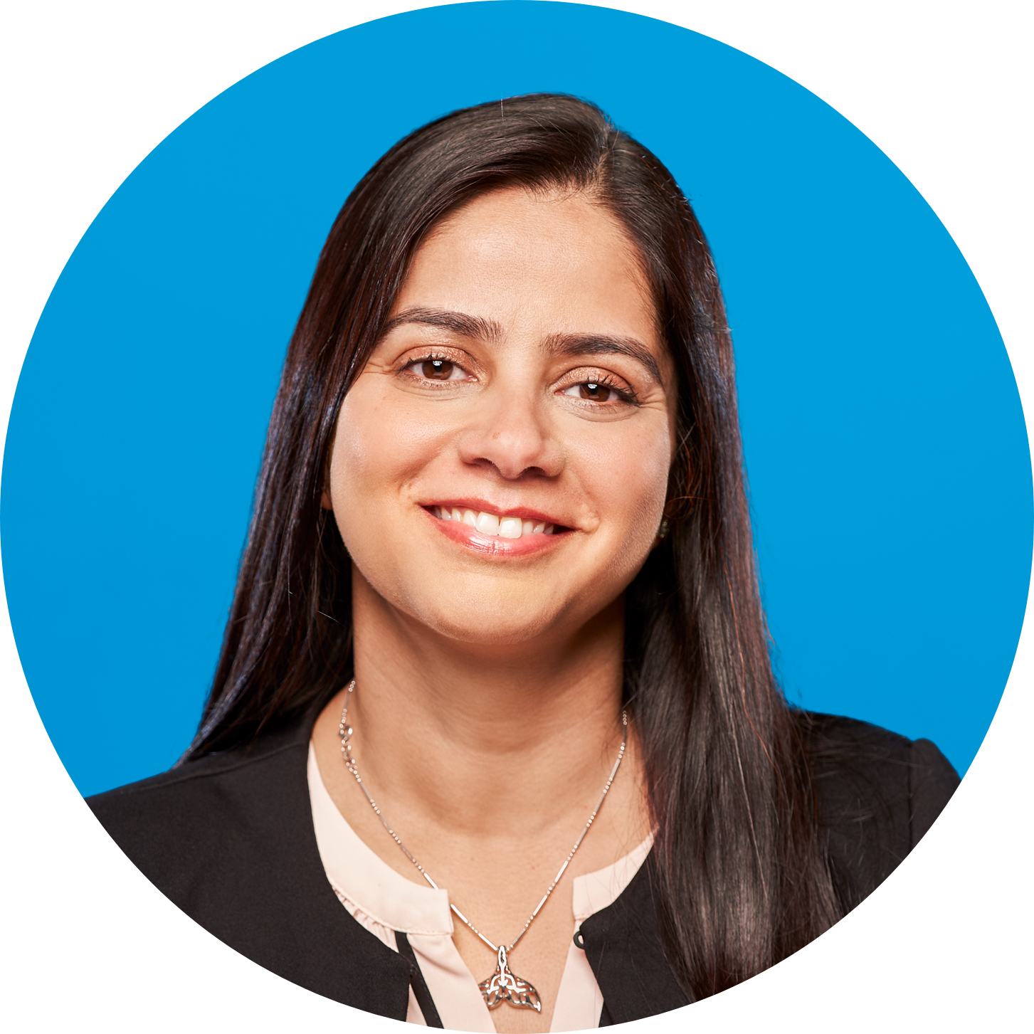 Headshot of smiling Madhavi wearing a black collarless jacket and gold necklace with pendant, against a deep blue background.