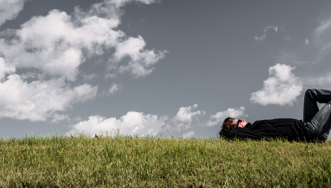 Man lying on his back on the grass, arms behind his head, looking up at a blue sky and puffy clouds