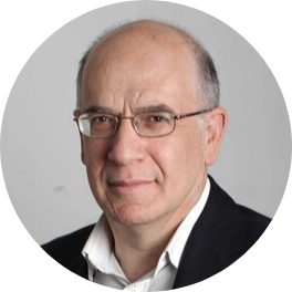 Headshot of Michael smiling against a grey background. He's wearing an open collar white shirt, black jacket and metal-framed glasses.