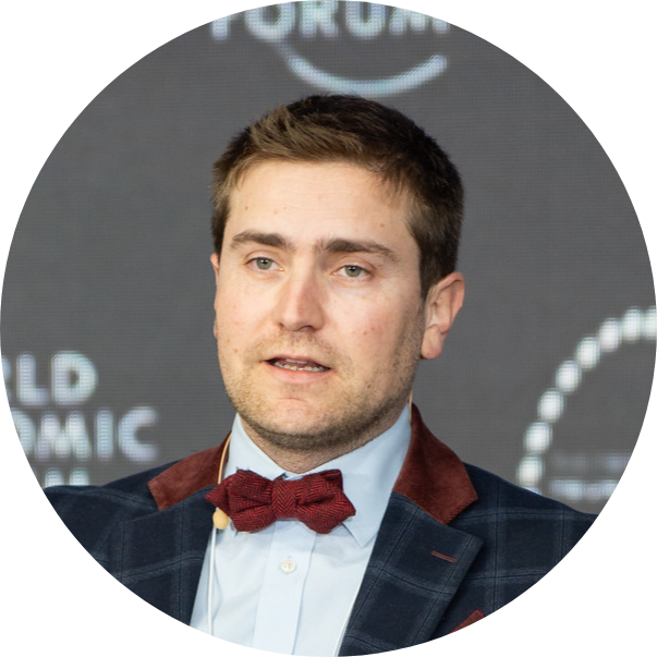 Headshot of Thomas speaking at the World Economic Forum, wearing a blue shirt, wine-colour bow tie and dark blazer.