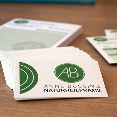 Angebote und Kosten Visitenkarten auf einem braunen Tisch. Die Karten zeigen das runde grüne Logo mit den Initialien AB. Darunter steht Anne Büssing Naturheilpraxis. im Hintergrund liegt unscharf der Rezeptblock und die Terminkarten.
