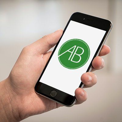 Kontakt Hand die ein Smartphone hält, welches das Logo von der Naturheilpraxis Anne Büssing zeigt. Das Logo ist ein runder grüner Kreis mit den Initialien AB