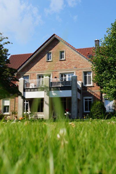 Praxis Anne Büssing Haus in dem die Praxis von Anne Büssing ist. Rotes Mauerwerk mit rotem Dach. An das Haus ist ein Balkon gebaut mit drei grauen Betonsäulen. Im Vordergrund unscharfer grüner Rasen. Das Haus wird von zwei Bäumen flankiert. Der Himmel ist blau und strahlend.