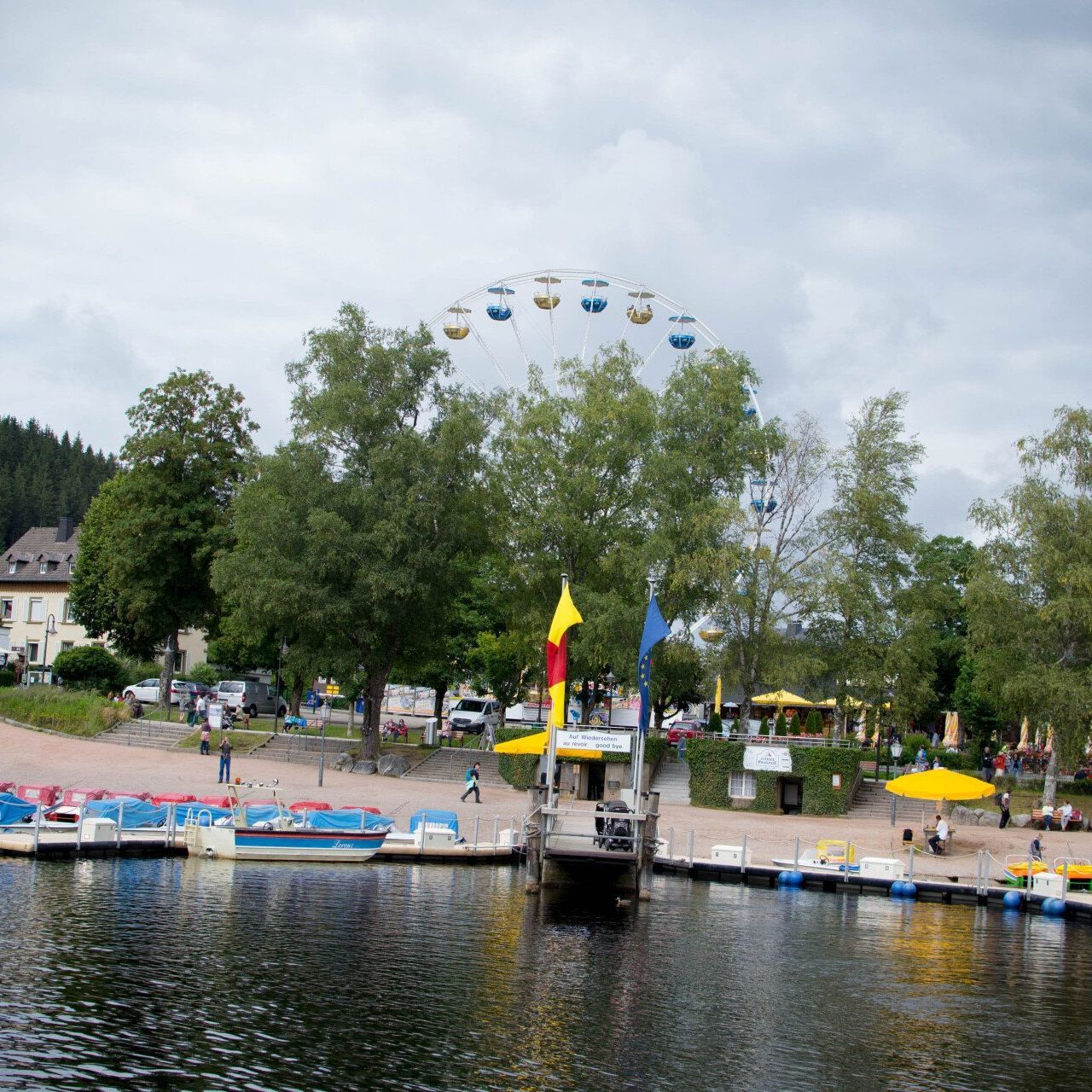 Familienurlaub Schwarzwald Titisee Blick auf das Ufer mit bunten Tretboten Strand Bäumen und einem Riesenrad