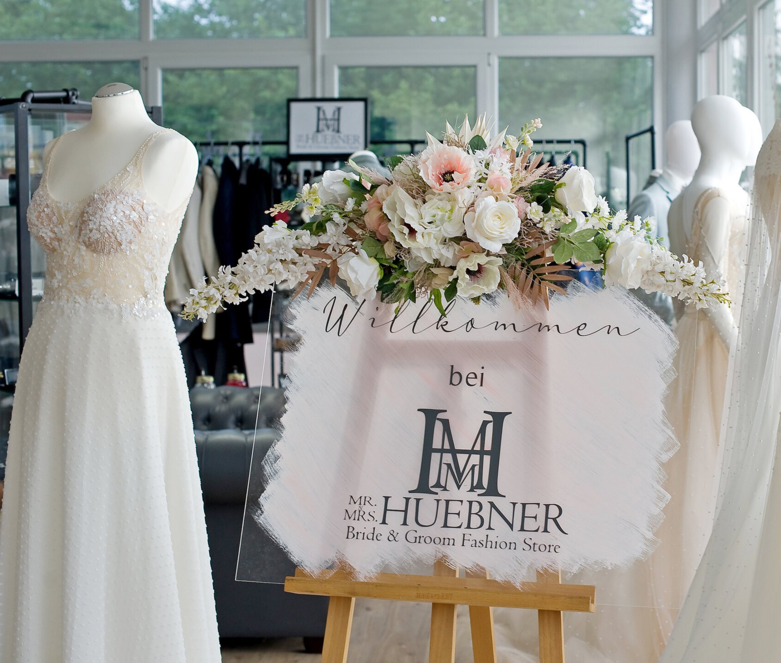 Ein stilvoll dekorierter Eingangsbereich im Brautmoden-Outlet Mr. & Mrs. Huebner in Gründau, nahe Wetzlar. Im Vordergrund steht ein Schild mit floraler Verzierung und der Aufschrift „Willkommen bei MR. & MRS. HUEBNER – Bride & Groom Fashion Store“. Darauf arrangiert ist ein opulenter Blumenstrauß mit weißen, roséfarbenen und grünen Elementen. Daneben steht eine Schneiderpuppe mit einem zarten Brautkleid aus Tüll und 3D-Blüten. Im Hintergrund sind weitere Braut- und Anzugmode sowie Schaufensterpuppen zu erkennen.