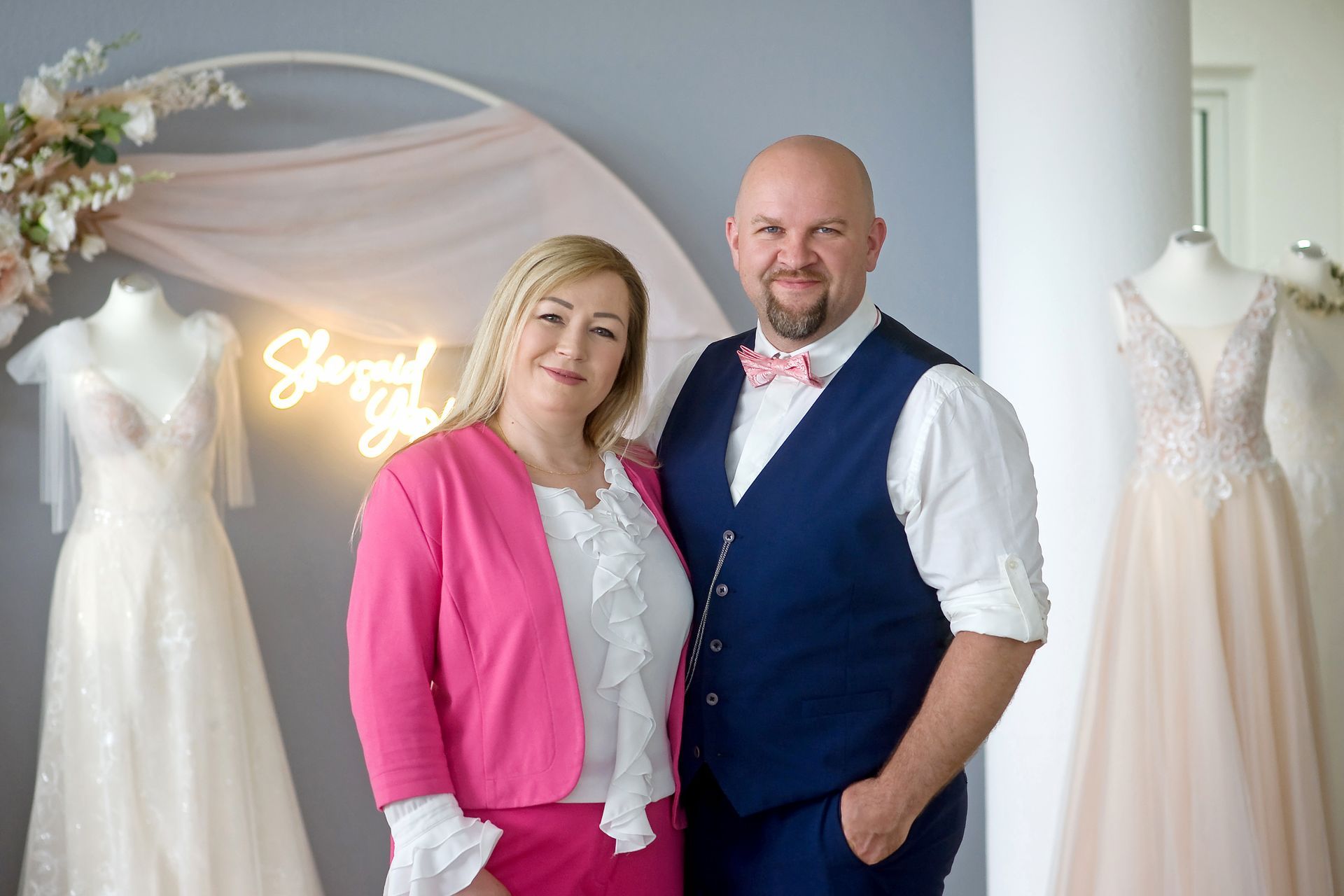 Das Bild zeit das Team von Mr & Mrs Huebner Brautmoden in Hessen im Main-Kinzig-Kreis. Im Hintergrund sieht man stilvolle Brautkleider. Vicotia Huebner trägt eine weiße Bluse mit einem zarte pinken Hosenanzug. Gerhard Huebner trägt ein weises Hemd mit einer blauen Weste-Hosen Kombi. Die beiden strahlen Freundlichkeit und Herzlickeit aus.