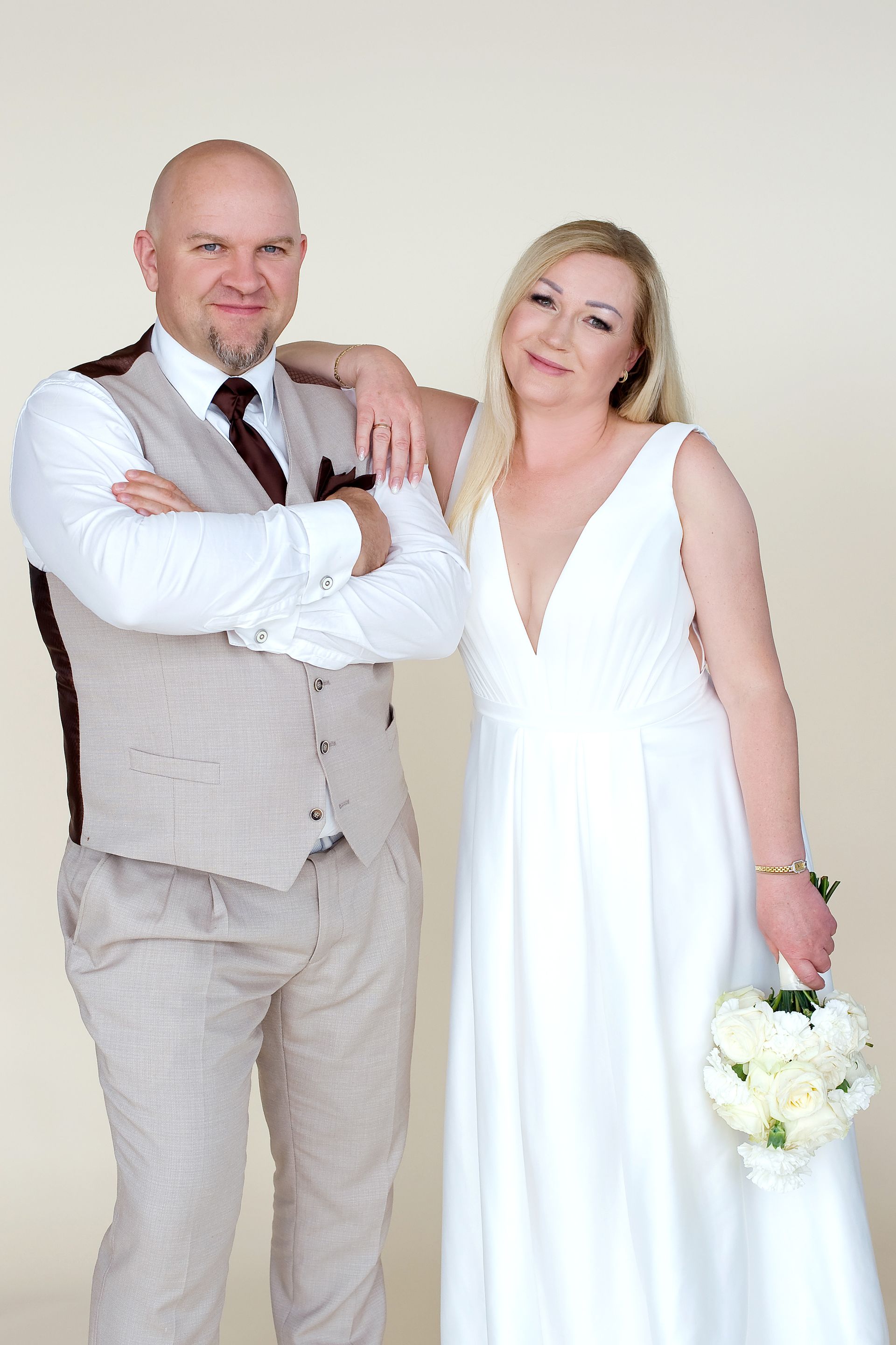Brautpaar in moderner Hochzeitsmode von Mr. & Mrs. Huebner aus Gründau. Der Bräutigam trägt einen eleganten sandfarbenen Hochzeitsanzug mit passender Weste, Krawatte und Einstecktuch. Die Braut präsentiert ein schlichtes, fließendes Brautkleid mit tiefem V-Ausschnitt und hält einen weißen Brautstrauß. Das Paar steht selbstbewusst nebeneinander und zeigt eine harmonisch aufeinander abgestimmte Hochzeitsgarderobe für moderne Hochzeiten. Individuelle Beratung für Brautkleider, Hochzeitsanzüge und perfekt abgestimmte Brautpaar-Outfits im Main-Kinzig-Kreis, nahe Frankfurt und Hanau.