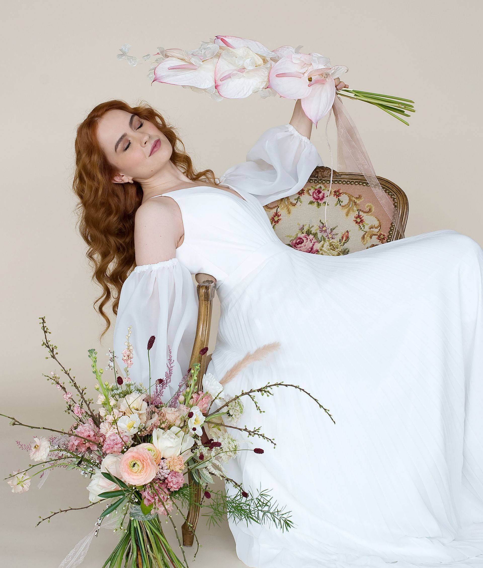 Eine junge Braut mit langen, gewellten, rotblonden Haaren sitzt entspannt auf einem antiken Stuhl mit floraler Polsterung. Sie trägt ein fließendes, modernes Brautkleid mit tiefem V-Ausschnitt und eleganten Puffärmeln aus transparentem Stoff. In der einen Hand hält sie einen Strauß aus zarten rosa Anthurien, in der anderen liegt ein üppiger Brautstrauß mit rosafarbenen, weißen und cremefarbenen Blüten. Die Szene wirkt ruhig, stilvoll und leicht verträumt – sie verkörpert moderne Brautmode im Vintage-Stil. Das Foto wurde im Brautmodengeschäft Mr & Mrs Huebner Brautmoden in der Nähe von Wetzlar aufgenommen und zeigt ein Hochzeitskleid aus der aktuellen Kollektion. Ideal für Bräute, die in Wetzlar ein einzigartiges Brautkleid suchen.