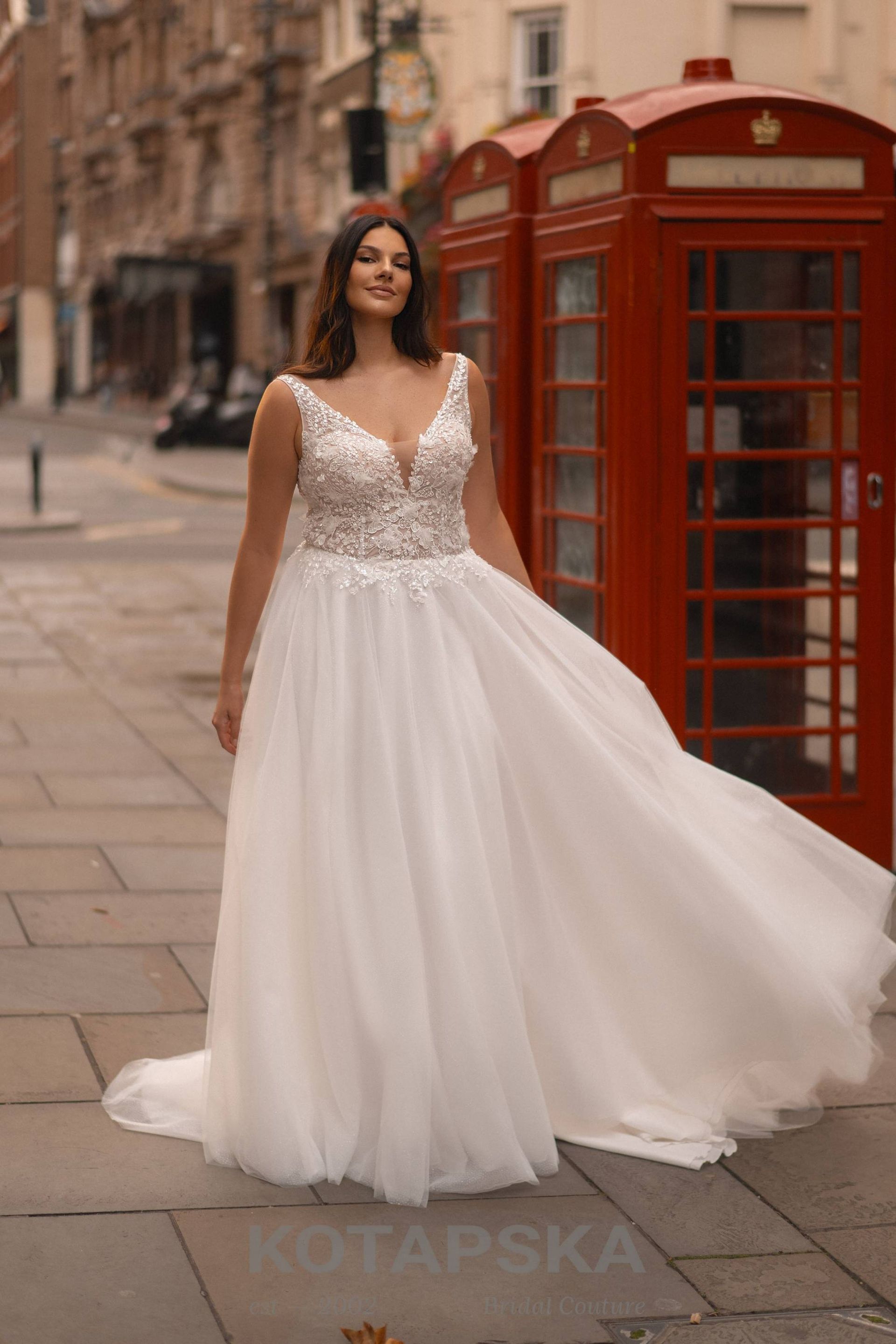 Die Braut trägt ein modernes Curvy-Brautkleid mit tiefem V-Ausschnitt und breiten, glitzernden Trägern. Das Oberteil ist mit floraler Spitze und Perlen verziert und betont das Dekolleté besonders elegant. Der weich fließende Tüllrock sorgt für Leichtigkeit und Bewegung. Die Braut schreitet selbstbewusst über eine gepflasterte Straße, im Hintergrund sind zwei klassische, rote Telefonzellen zu sehen.
Dieses Curvy-Brautkleid in Übergröße ist erhältlich bei Mr. & Mrs. Hübner Brautmoden nahe Hanau.