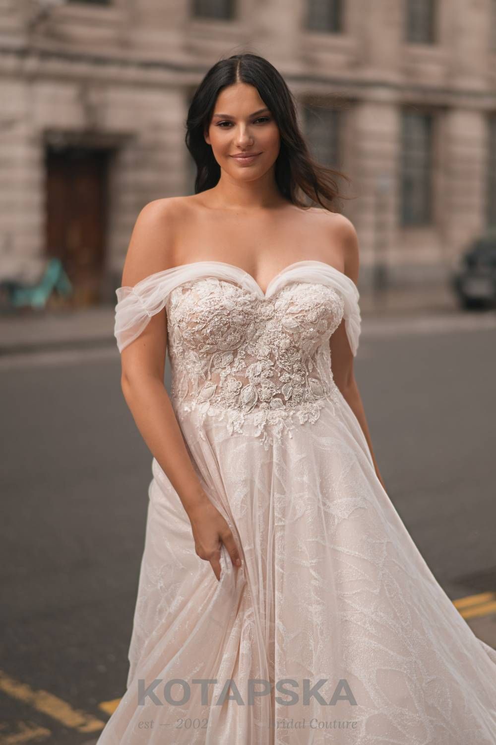 Die Braut trägt ein sinnliches Curvy-Brautkleid mit schulterfreier Corsage und Herzausschnitt. Zarte Tüllärmel umschmeicheln die Oberarme, während das Spitzenoberteil mit floralen Applikationen und leicht transparentem Einsatz verziert ist. Der weich fließende Rock rundet den femininen Look ab. Die Braut steht auf einer städtischen Straße vor einem historischen Gebäude und lächelt sanft in die Kamera.
Dieses Curvy-Brautkleid in Übergröße ist erhältlich bei Mr. & Mrs. Hübner Brautmoden nahe Birstein