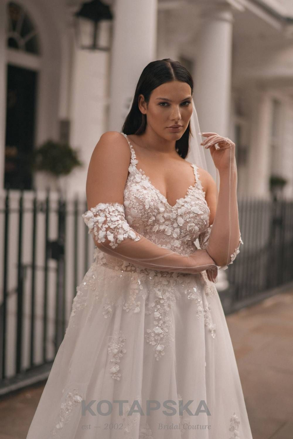 Die Braut trägt ein detailverliebtes Curvy-Brautkleid mit schmalen Trägern und aufwendig besticktem Spitzenoberteil. Florale 3D-Applikationen und zarte Glitzerdetails verleihen dem Kleid eine luxuriöse Ausstrahlung. Die Ärmel aus transparenter Spitze sind locker über die Oberarme gezogen und sorgen für einen romantisch-verspielten Look. Die Braut hält den Schleier in der Hand und steht vor einem weißen Säulengang mit schwarzem Geländer.
Dieses Curvy-Brautkleid in Übergröße ist erhältlich bei Mr. & Mrs. Hübner Brautmoden nahe Gießen.