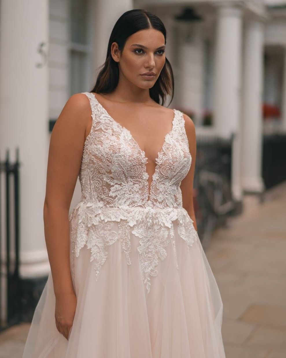 Eine junge Braut trägt ein modernes Curvy Brautkleid mit tiefem V-Ausschnitt, transparentem Oberteil und detailreicher floraler Spitze. Das Plus Size Brautkleid betont das Dekolleté und die Taille auf elegante Weise. Die Aufnahme zeigt sie im Halbprofil vor einer weißen Häuserfassade mit dezentem Stadthintergrund. Dieses Modell steht für stilvolle Curvy Brautmode, Plus Size Brautmode und Brautmode in Übergrößen. Erhältlich im Brautmodengeschäft Mr. & Mrs. Hübner in Gründau in der Nähe von Wächtersbach