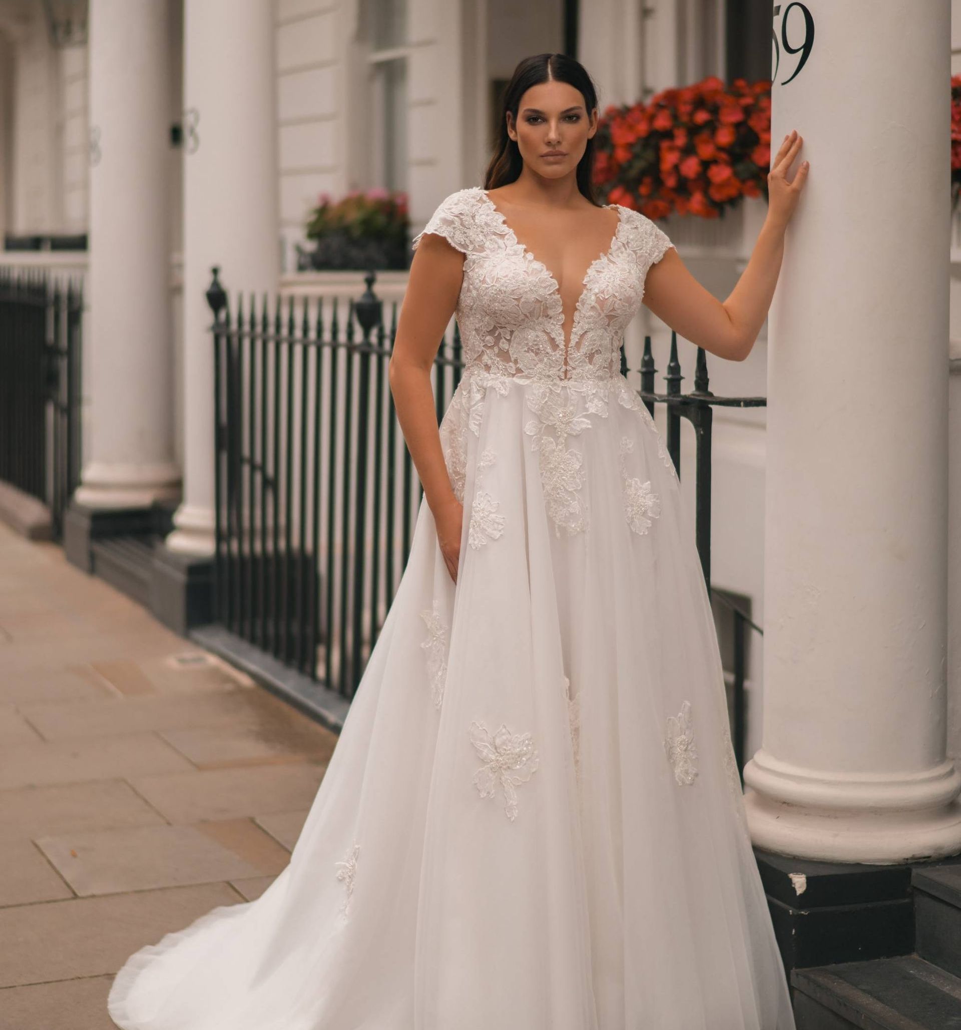 Eine junge Braut trägt ein elegantes Curvy-Brautkleid mit tiefem V-Ausschnitt, floralen Spitzenapplikationen und leicht angeschnittenen Ärmeln. Das Plus Size Brautkleid in A-Linie betont die Figur sanft und fällt weich bis zum Boden. Der Hintergrund zeigt eine weiße Stadtfassade mit schwarzen Geländern und blühenden roten Blumen. Dieses Modell steht für moderne Curvy Brautmode, Plus Size Brautmode und Brautmode in Übergrößen. Erhältlich im Brautmodengeschäft Mr. & Mrs. Hübner in Gründau, in der Nähe von Leidersbach