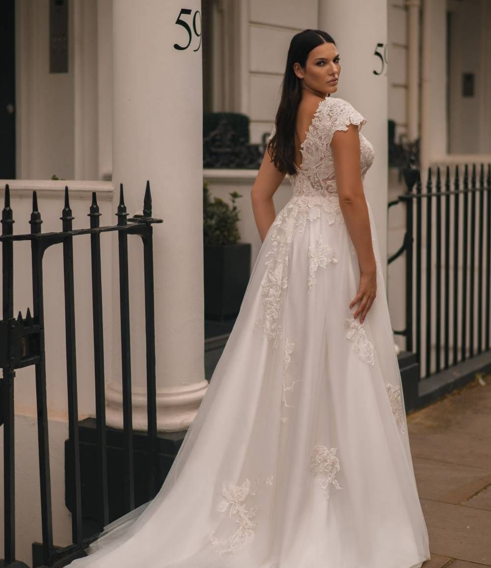 Eine junge Braut steht mit dem Rücken zur Kamera und trägt ein elegantes Curvy Brautkleid mit zarter Spitze, angeschnittenen Ärmeln und langem fließenden Rock. Das Rücken-Dekolleté ist tief geschnitten und unterstreicht die feminine Silhouette. Die Aufnahme wurde vor einer weißen Hausfassade mit schmiedeeisernen Zäunen gemacht. Das Brautkleid ist Teil der Curvy Brautmode, Plus Size Brautmode und Brautmode in Übergrößen bei Mr. & Mrs. Hübner in Gründau in der Nähe von Sinntal