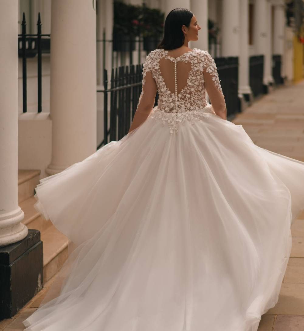 Die Braut trägt ein romantisches Curvy-Brautkleid mit weitem Tüllrock und aufwendig gearbeitetem Spitzenoberteil. Das Oberteil hat halblange Ärmel aus transparenter Spitze mit 3D-Blüten und ist im Rücken mit einem eleganten Cut-out versehen. Der weich fallende Rock verleiht dem Kleid eine traumhaft leichte Bewegung. Die Braut hat dunkles, langes Haar und wird von hinten gezeigt, während sie einen städtischen, säulengesäumten Gehweg entlangschreitet.
Dieses Curvy-Brautkleid in Übergröße ist erhältlich bei Mr. & Mrs. Hübner Brautmoden nahe Frankfurt.