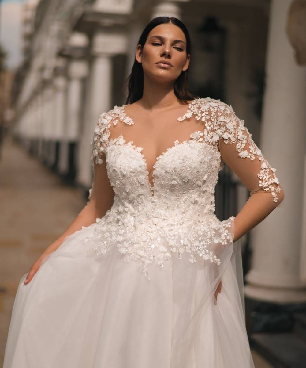 Eine junge Braut trägt ein elegantes Curvy Brautkleid mit transparenten langen Ärmeln aus floraler 3D-Spitze und einem tiefen Herzausschnitt. Das Oberteil ist mit filigranen Blütenapplikationen verziert, der weiche Tüllrock fällt fließend bis zum Boden. Die Aufnahme zeigt sie im Sonnenlicht vor einer Säulenfassade in einer städtischen Kulisse. Dieses Brautkleid ist Teil der Curvy Brautmode, Plus Size Brautmode und Brautmode in Übergrößen bei Mr. & Mrs. Hübner in Gründau,  in der Nähe von Aschaffenburg
