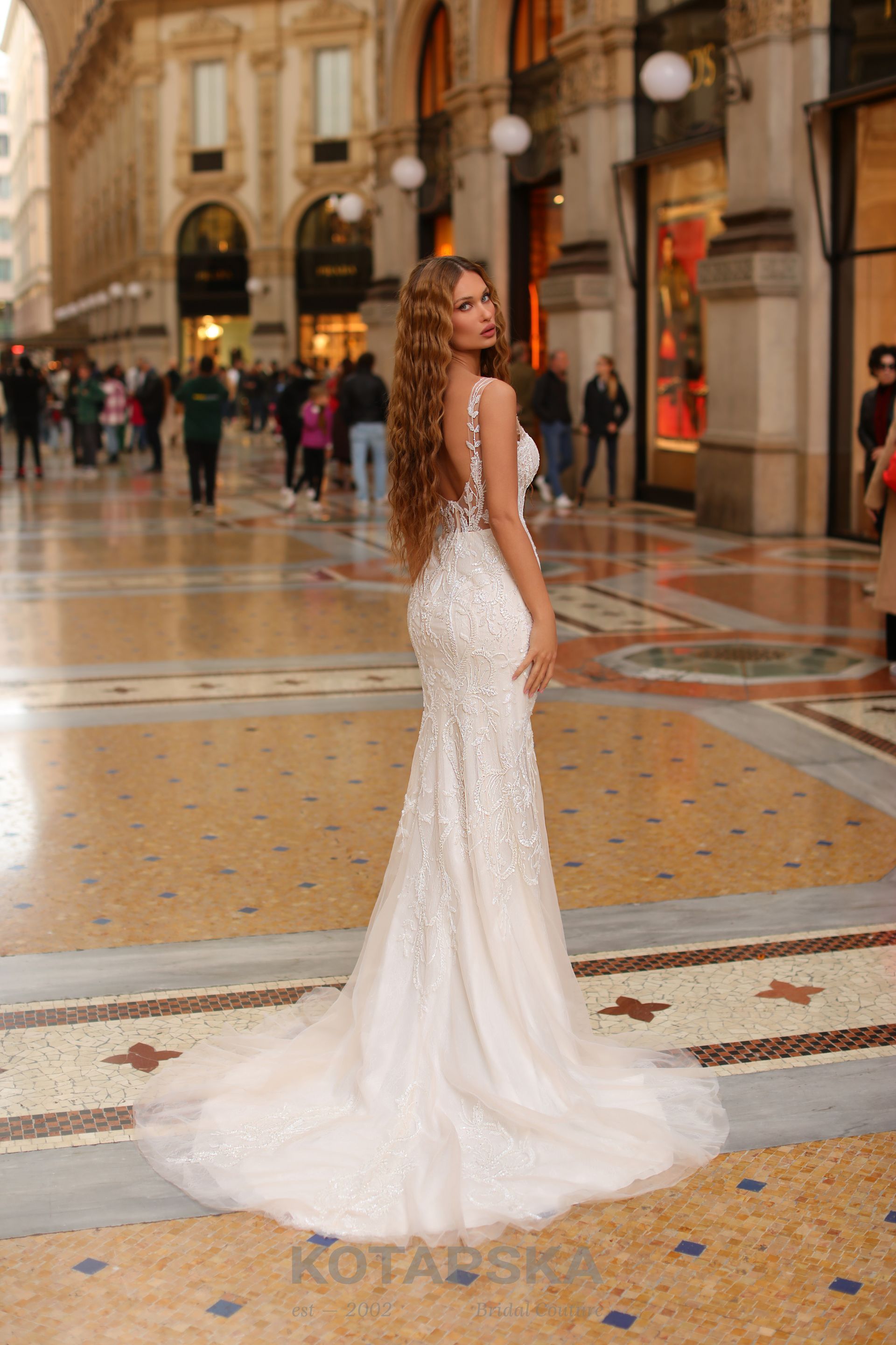 Die Braut trägt ein figurbetontes, elegantes Brautkleid im Fit-and-Flare-Stil. Das Kleid ist aus feiner Spitze gefertigt, hat einen tiefen Rückenausschnitt und transparente Träger mit Spitzenapplikationen. Der Schnitt betont die Silhouette der Braut und läuft nach unten hin leicht aus. Die Braut hat lange, gewellte Haare und blickt über die Schulter zurück. Sie steht in einer prunkvollen Einkaufspassage mit Mosaikboden und Arkaden, umgeben von Passanten im Hintergrund.
Dieses Brautkleid ist erhältlich bei Mr. & Mrs. Hübner Brautmoden nahe  Birstein