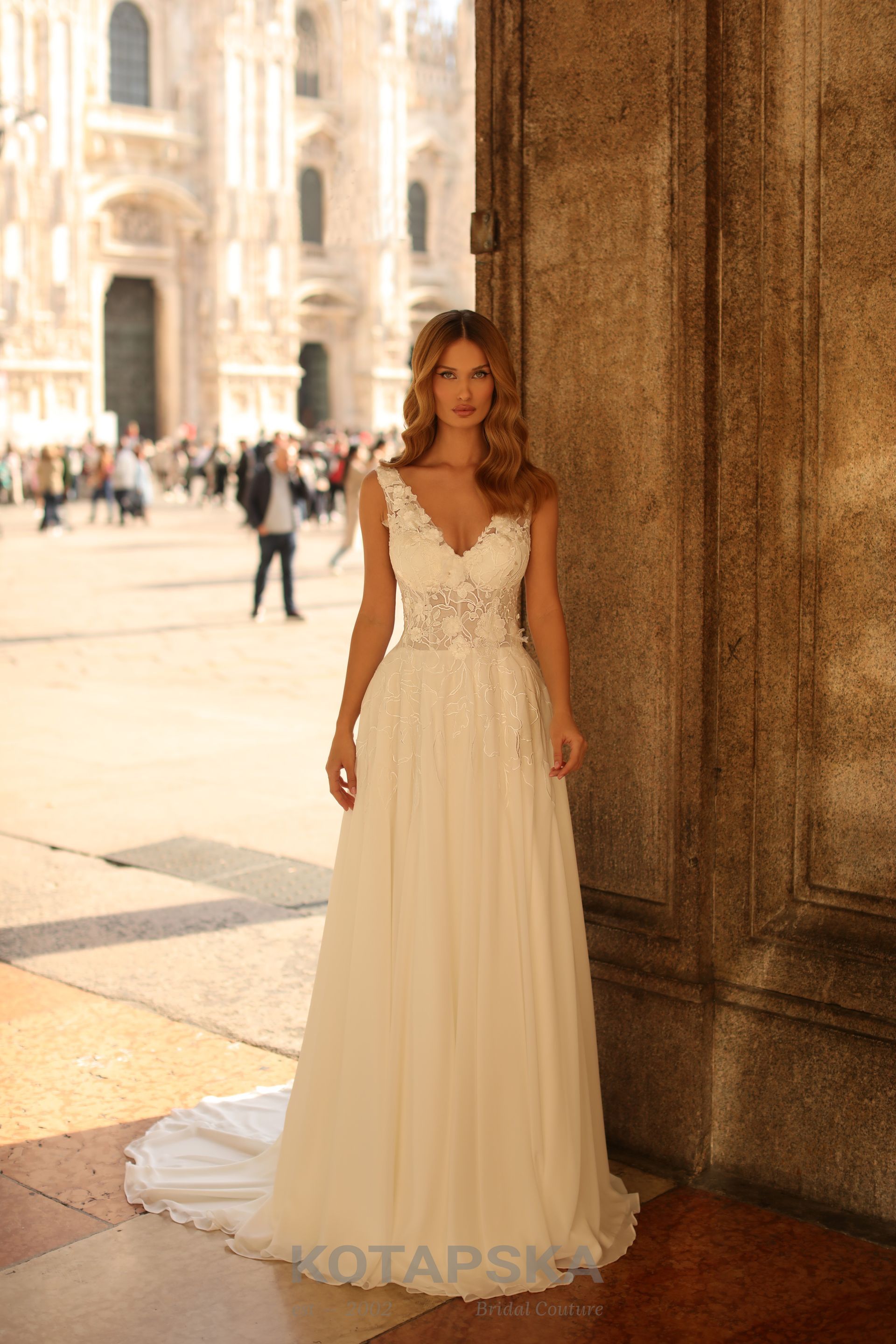 Die Braut trägt ein zartes, fließendes Brautkleid im A-Linien-Stil mit leichtem V-Ausschnitt. Das Oberteil ist mit feiner Spitze verziert und geht elegant in einen schlichten, weich fallenden Rock über. Die Träger des Kleides sind ebenfalls mit Spitzenapplikationen geschmückt. Die Braut hat offene, sanft gewellte Haare und steht im Halbschatten neben einer historischen Steinsäule. Im Hintergrund ist ein belebter Platz mit Menschen und klassischer Architektur zu sehen.
Dieses Brautkleid ist erhältlich bei Mr. & Mrs. Hübner Brautmoden nahe  Aschaffenburg.