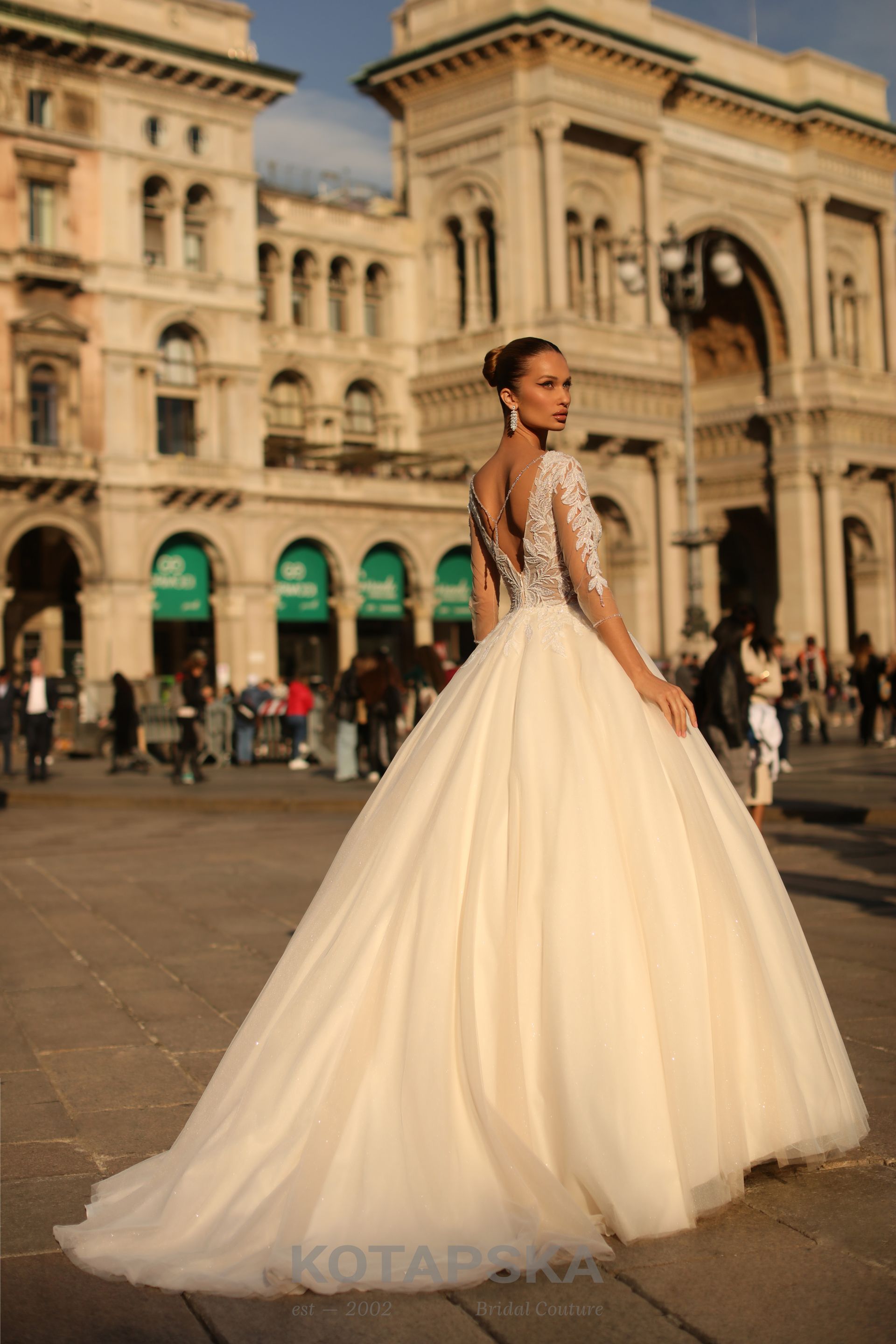 Die Braut trägt ein opulentes Prinzessinnenkleid mit weit ausgestelltem Rock und langer Schleppe. Das Oberteil besticht durch einen tiefen Rückenausschnitt und lange, transparente Spitzenärmel, die für eine edle und klassische Note sorgen. Die Haare der Braut sind elegant hochgesteckt. Sie steht auf einem historischen Platz vor einem imposanten Gebäude mit Bögen und Fassadendetails, während sich im Hintergrund Menschen tummeln.
Dieses Brautkleid ist erhältlich bei Mr. & Mrs. Hübner Brautmoden nahe Langenselbold.