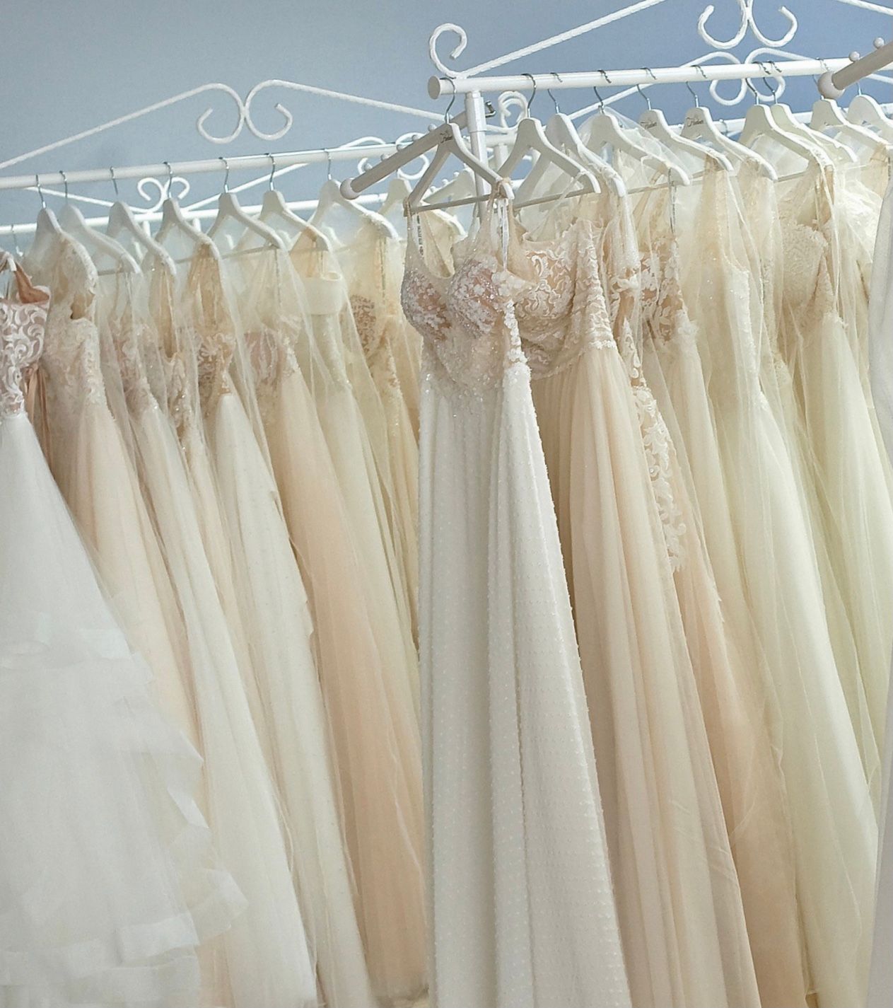Eine Reihe eleganter Brautkleider hängt dicht nebeneinander auf weißen Metallbügeln an einer dekorativen weißen Stange. Die Kleider sind in sanften Creme-, Champagner- und Blushtönen gehalten und zeichnen sich durch zarte Spitze, Tüll und glitzernde Details aus. Im Fokus steht ein fließendes Brautkleid mit transparenten Trägern und floraler Stickerei im Oberteil.
Aufgenommen im Brautmoden-Outlet Gründau nahe Fulda