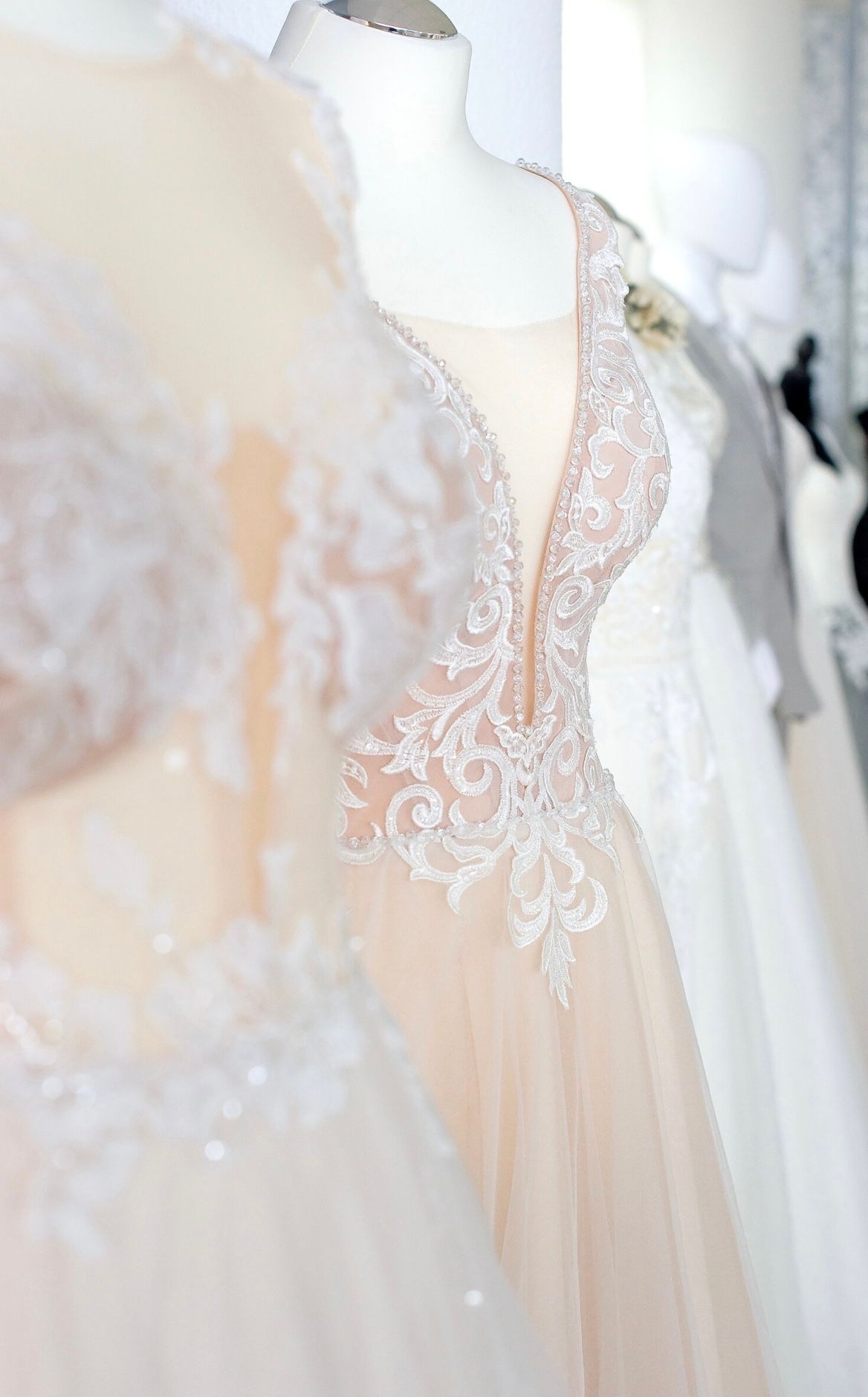 Im Vordergrund ist ein Brautkleid auf einer Schneiderpuppe zu sehen, das durch ein tiefes V-Dekolleté mit feiner, weißer Stickerei auf transparentem Nude-Tüll besticht. Die floralen Ornamente und glitzernden Details verleihen dem Kleid eine besonders elegante und moderne Ausstrahlung. Im Hintergrund sind weitere Brautkleider auf Puppen zu erkennen.
Aufgenommen im Brautmoden-Outlet Gründau nahe Fulda