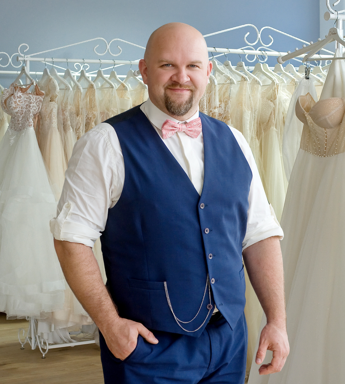 Inhaber von Mr. & Mrs. Huebner Brautmode in Gründau präsentiert sich in einem eleganten blauen Anzug mit Fliege vor einer Auswahl hochwertiger Brautkleider. Im Hintergrund befinden sich zahlreiche moderne Hochzeitskleider in verschiedenen Stilrichtungen. Das Bild steht für persönliche Beratung, individuelle Betreuung und exklusive Brautmode für Bräute und Bräutigame im Main-Kinzig-Kreis, nahe Frankfurt, Hanau und Fulda. Spezialist für Brautkleider, Standesamtkleider, Hochzeitsanzüge und perfekt abgestimmte Brautpaar-Outfits.
