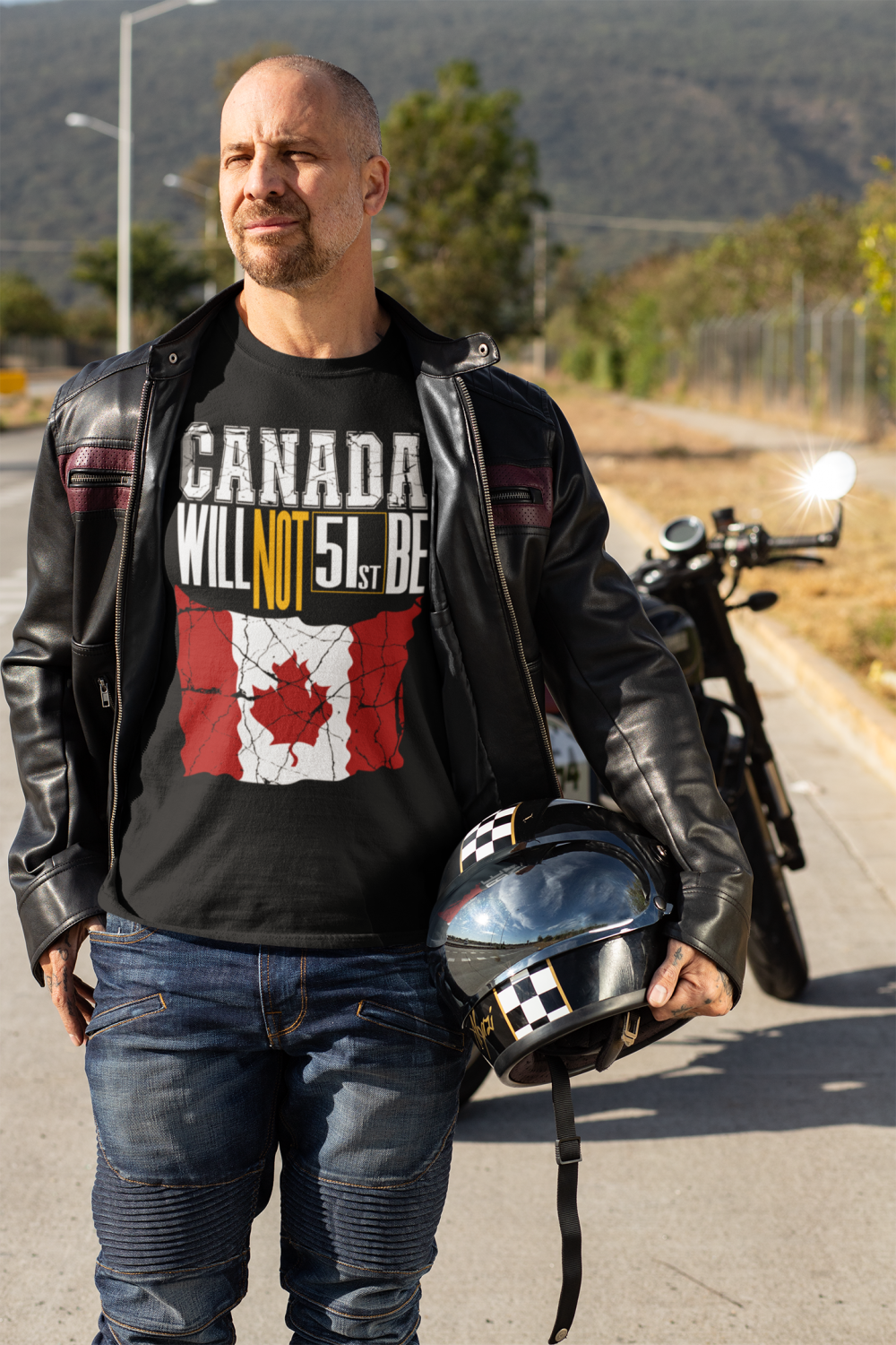 A man wearing a black Canada ‘Will Not Be 51st’ political graphic T-shirt, standing near a motorcycle in an outdoor setting.