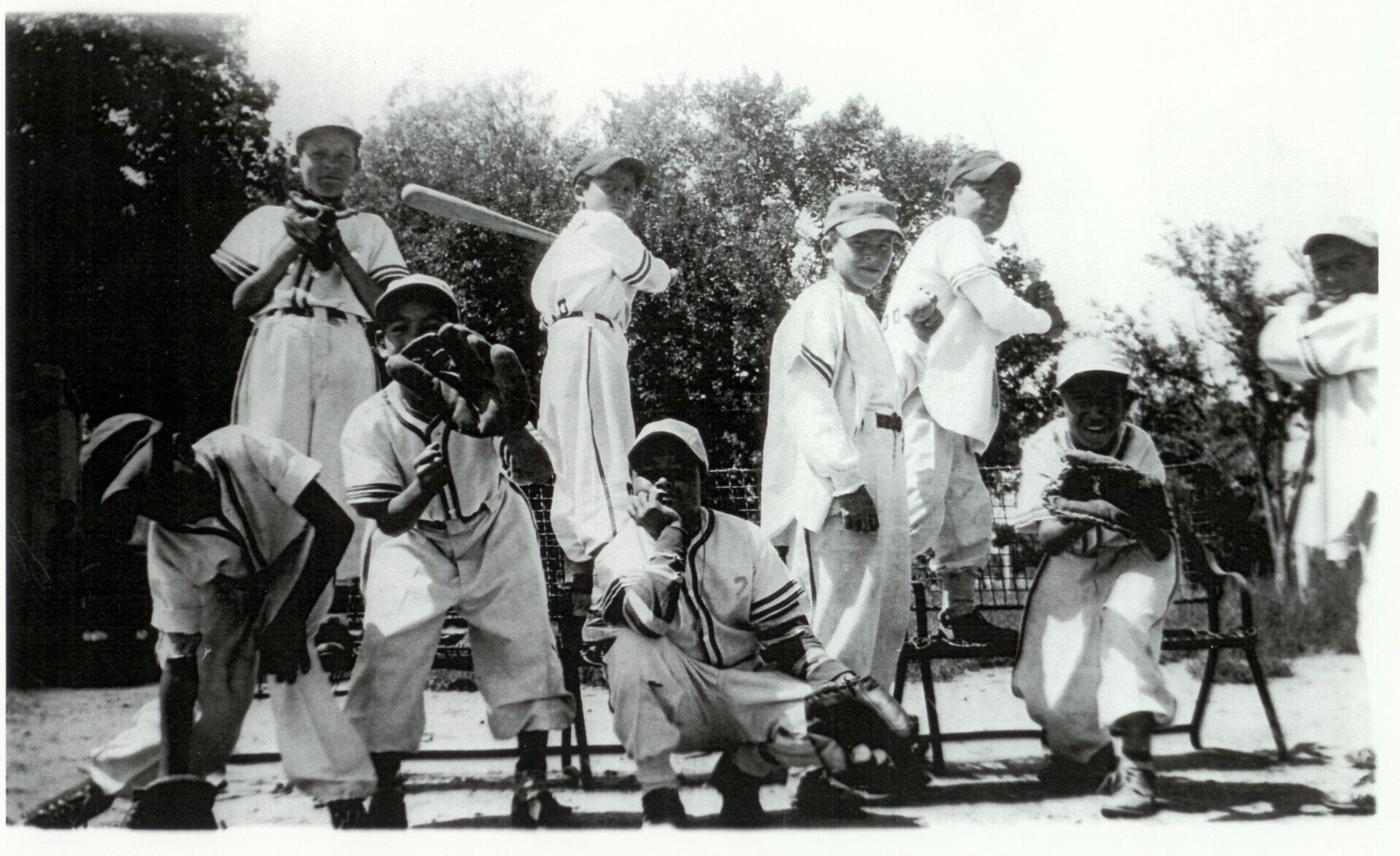 Black and white photograph of nine young boys in baseball uniforms, some holding bats and gloves, posing for a team photo outdoors with trees in the background.