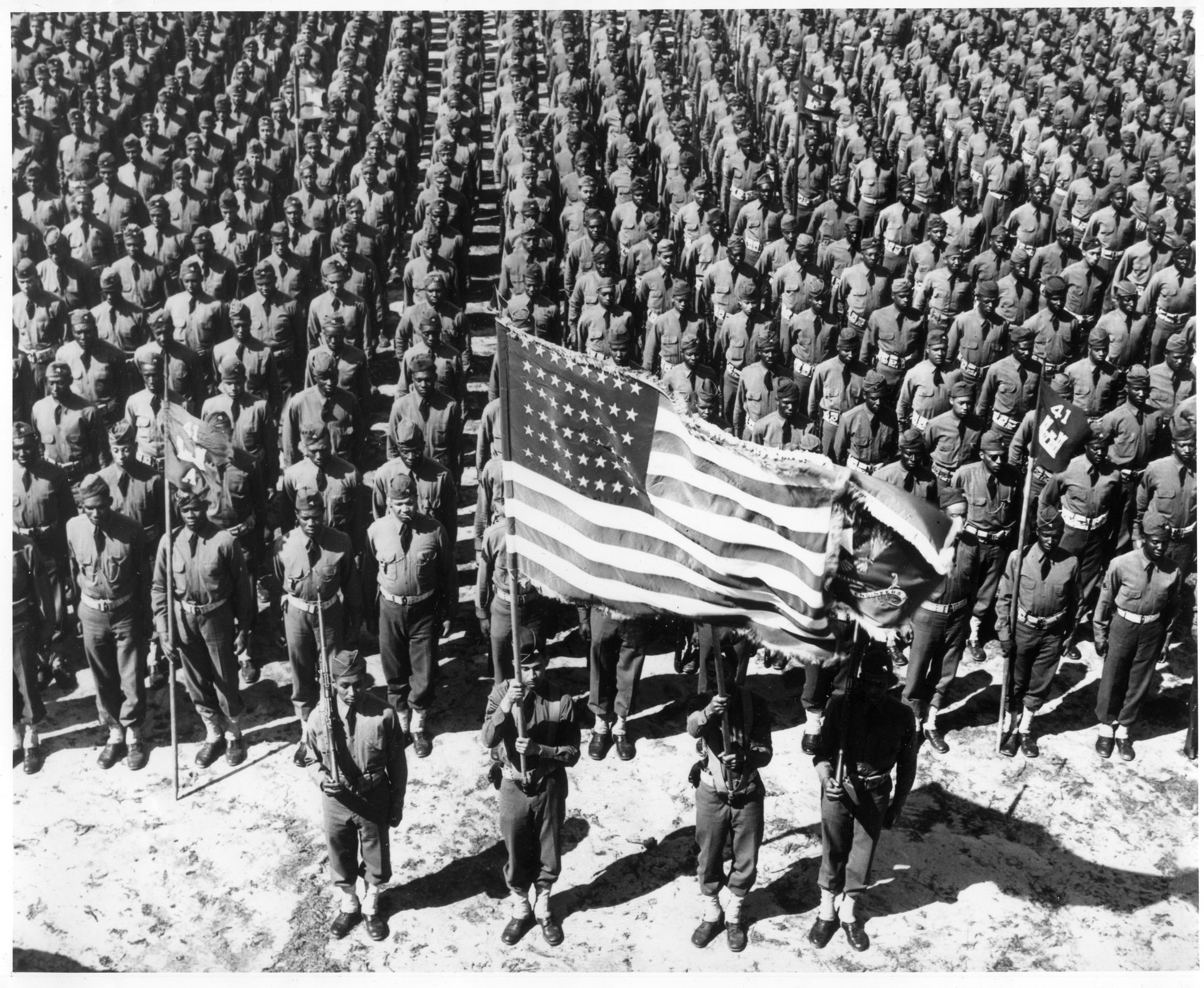 Hundreds of Black soldiers in uniform stand in formation on a dirt field, with an American flag and banner held in the foreground.