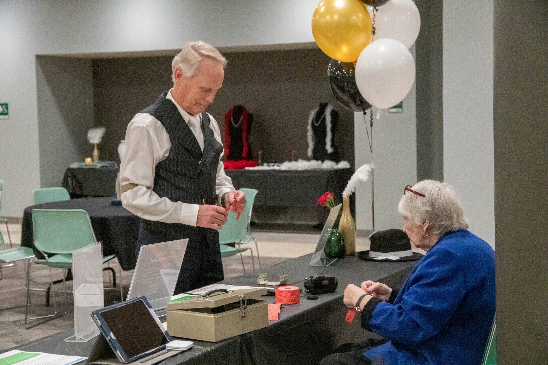 Man in pinstripe vest and woman in blue jacket at a black draped table with balloons and tickets.