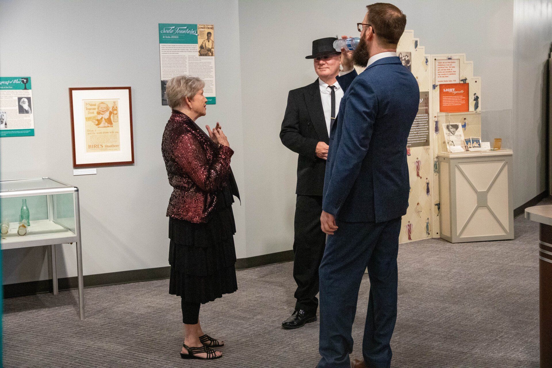 Three people converse in a museum exhibit with framed historical beverage advertisements and display cases.