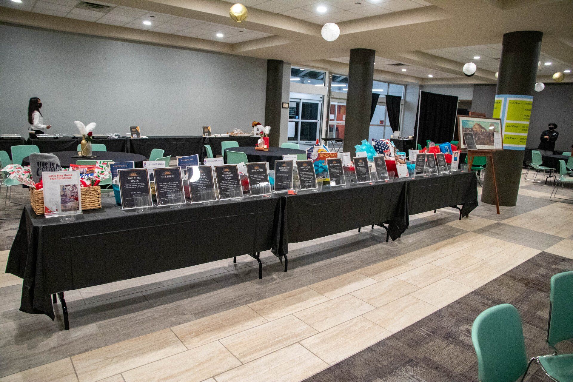 Silent auction display with numerous items and descriptions on black-covered tables in a well-lit room.