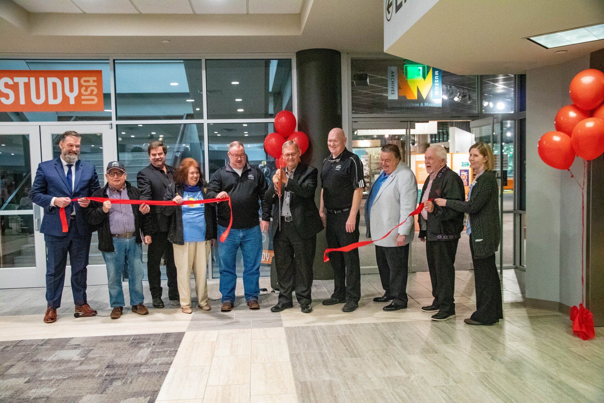 Group of people holding a red ribbon, celebrating grand opening, man in center snips ribbon with oversized scissors.