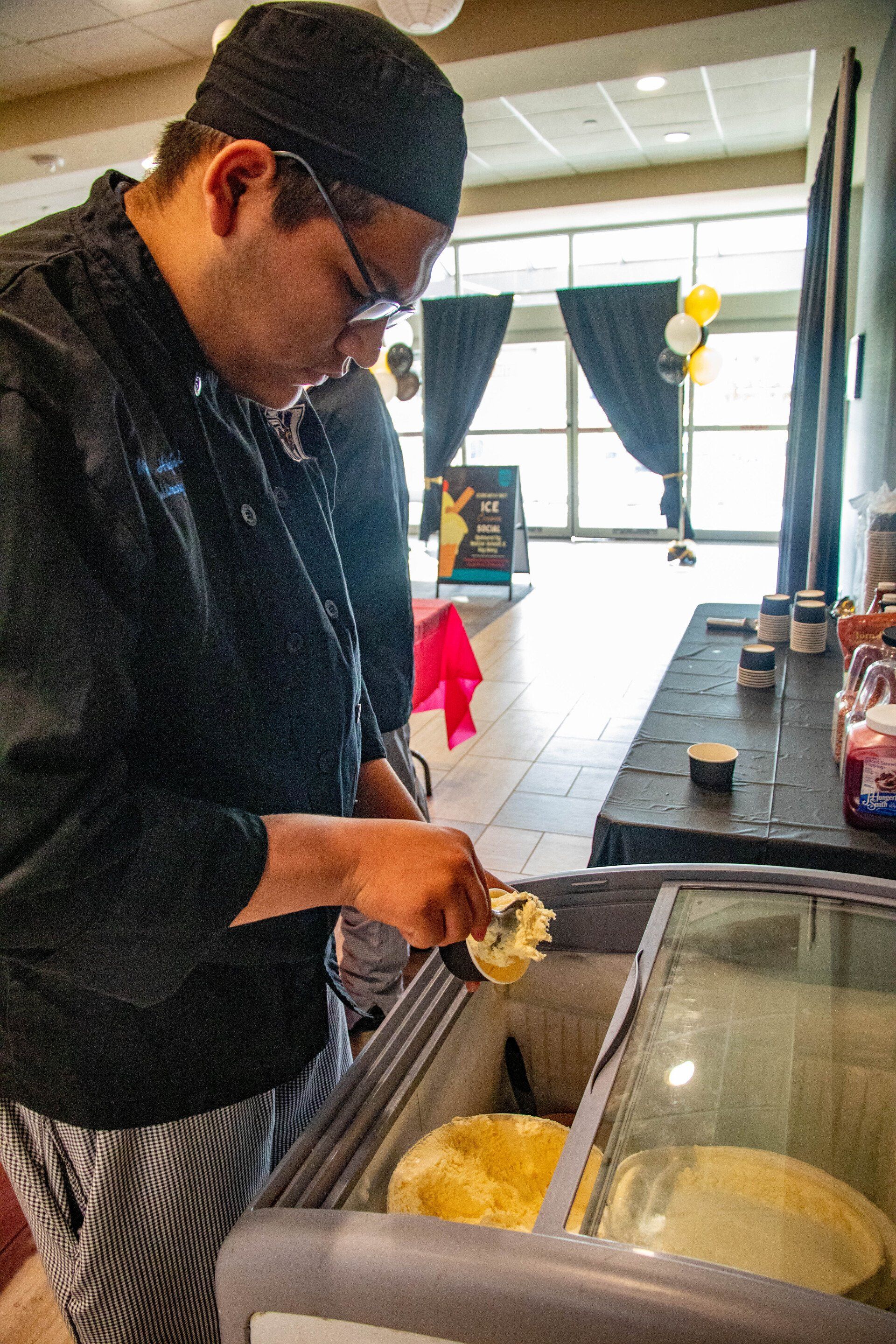 Person in chef coat scooping vanilla ice cream from a freezer into a cup at an ice cream social.