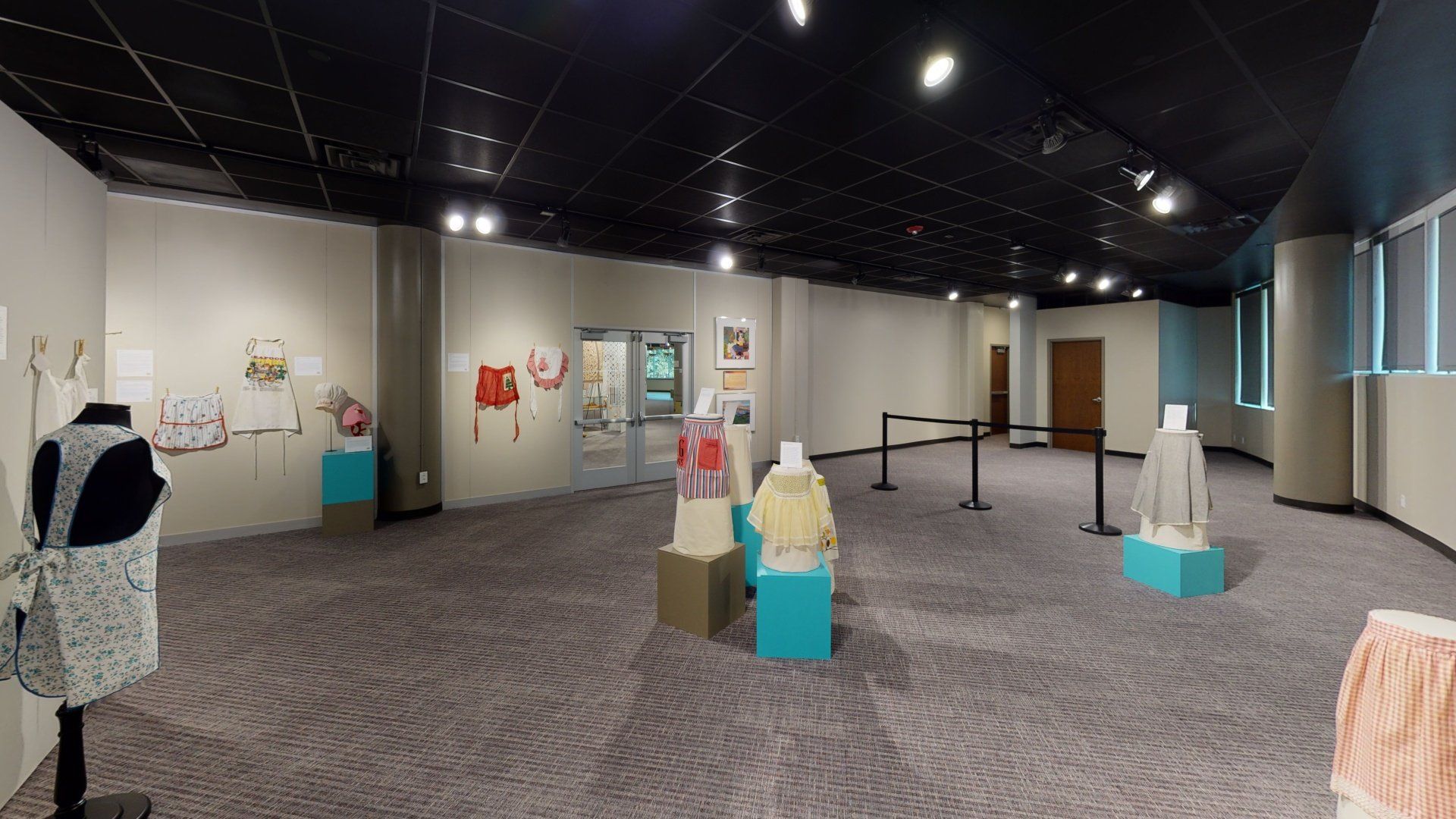 Exhibition hall with aprons displayed on mannequins, walls, and in cases, with windows on the right providing natural light.
