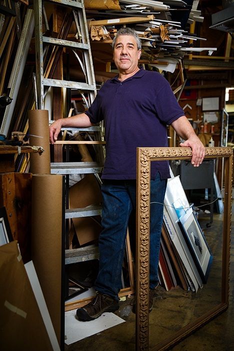 Man holding an ornate gold picture frame in a busy workshop surrounded by framing supplies.