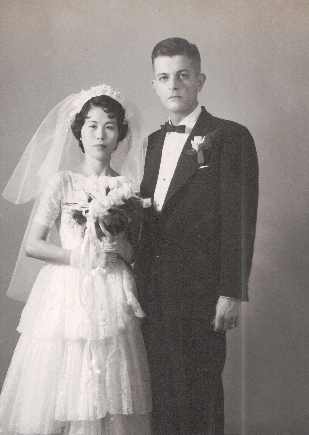 Black and white portrait of a bride in a lace dress with veil and bouquet, standing next to a groom in a tuxedo.