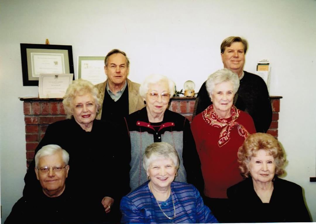 Group of seven adults, four women and three men, posing in front of a fireplace with framed certificates and a globe.
