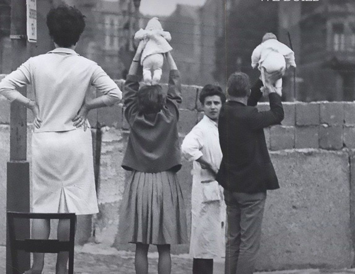 A black and white image depicts a group of people standing on one side of a wall, holding babies up over the top.