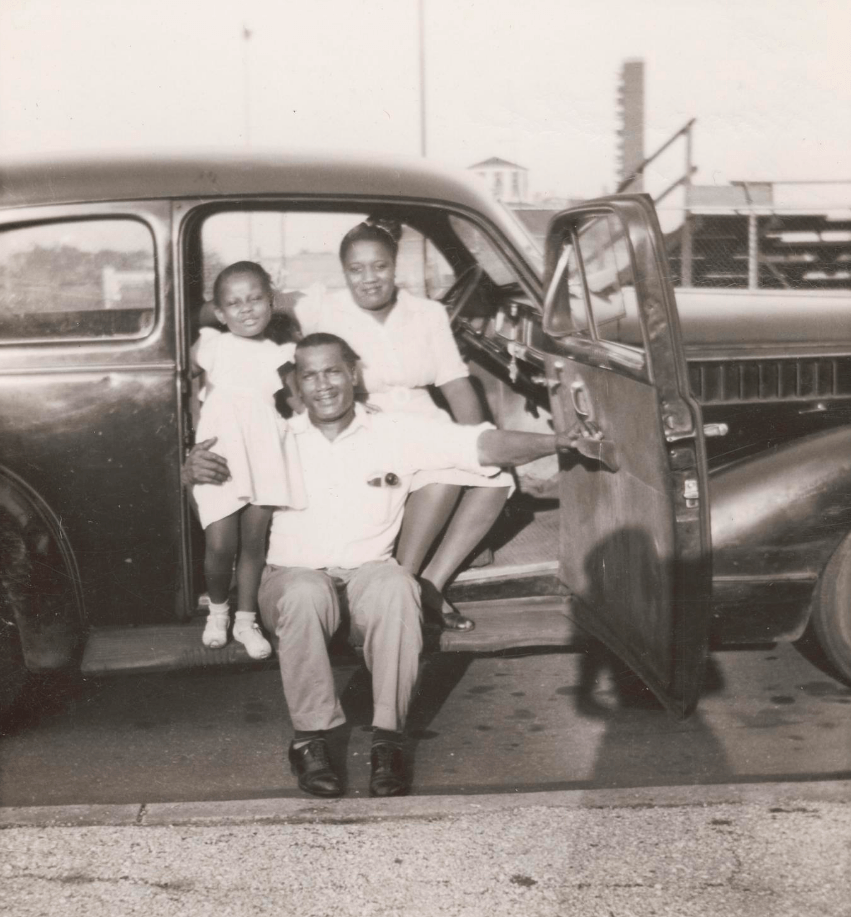 African American family of three in a classic car, smiling. Man holds child, woman sits in back. Vintage sedan.