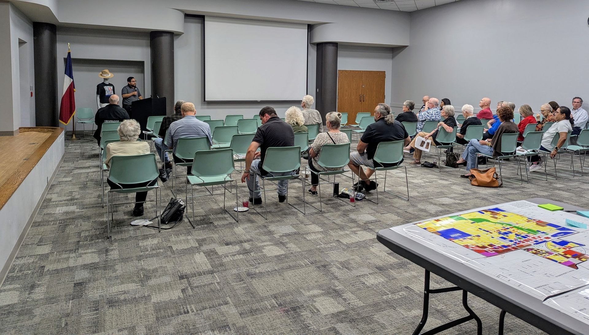 Speaker addresses an audience at a public meeting with a Texas flag and large screen in the background. A map is on a table.