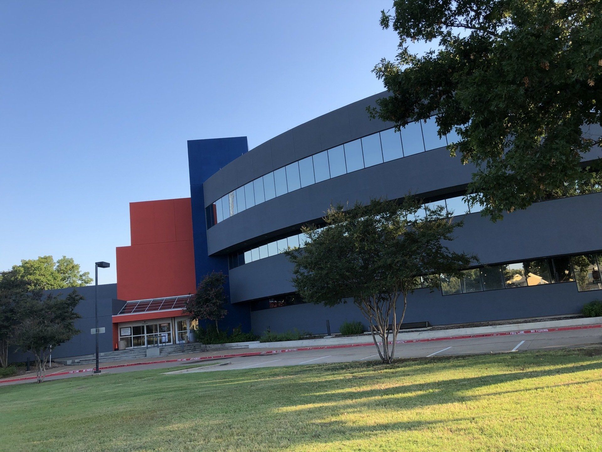 Modern building with curved gray facade, rectangular windows, and sections of red and blue, with grass and trees.
