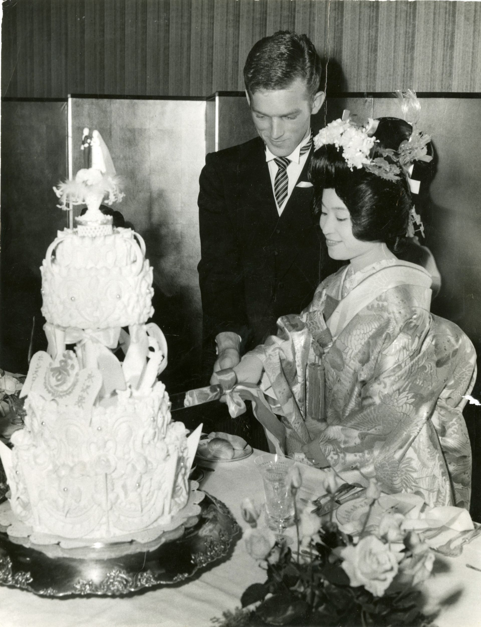 Man in tuxedo and woman in kimono cutting a three-tier wedding cake with a bride and groom figurine and decorative pearls.